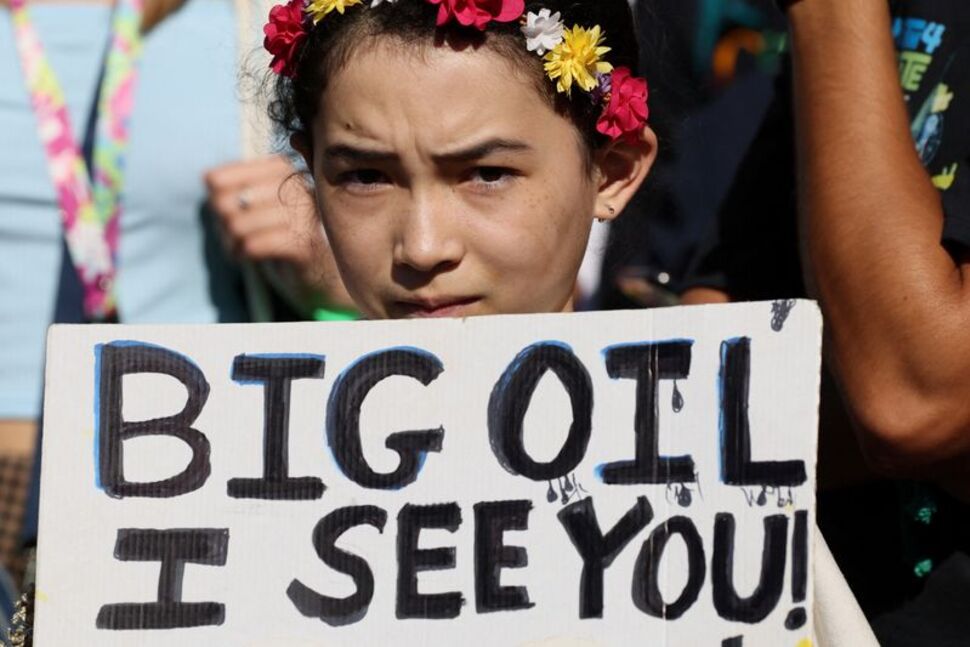 Girl with a floral headdress and a determined expression, carrying a sign reading 'BIG OIL I SEE YOU.
